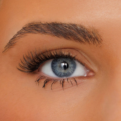 Close-up of a blue-gray eye with long black eyelashes, a defined eyebrow, and smooth warm-toned skin.