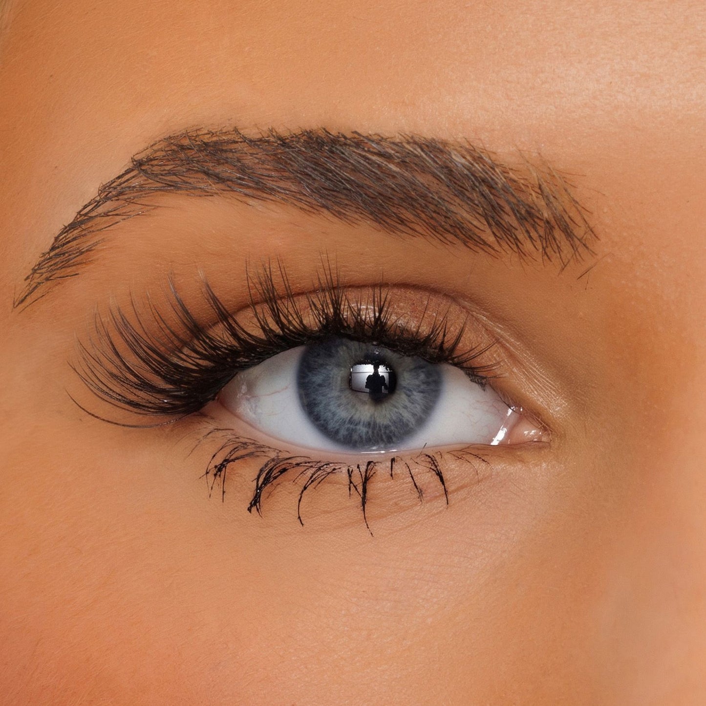 Close-up of a person's eye with long, dark, curled false eyelashes and a neatly shaped dark brown eyebrow on warm tan skin.