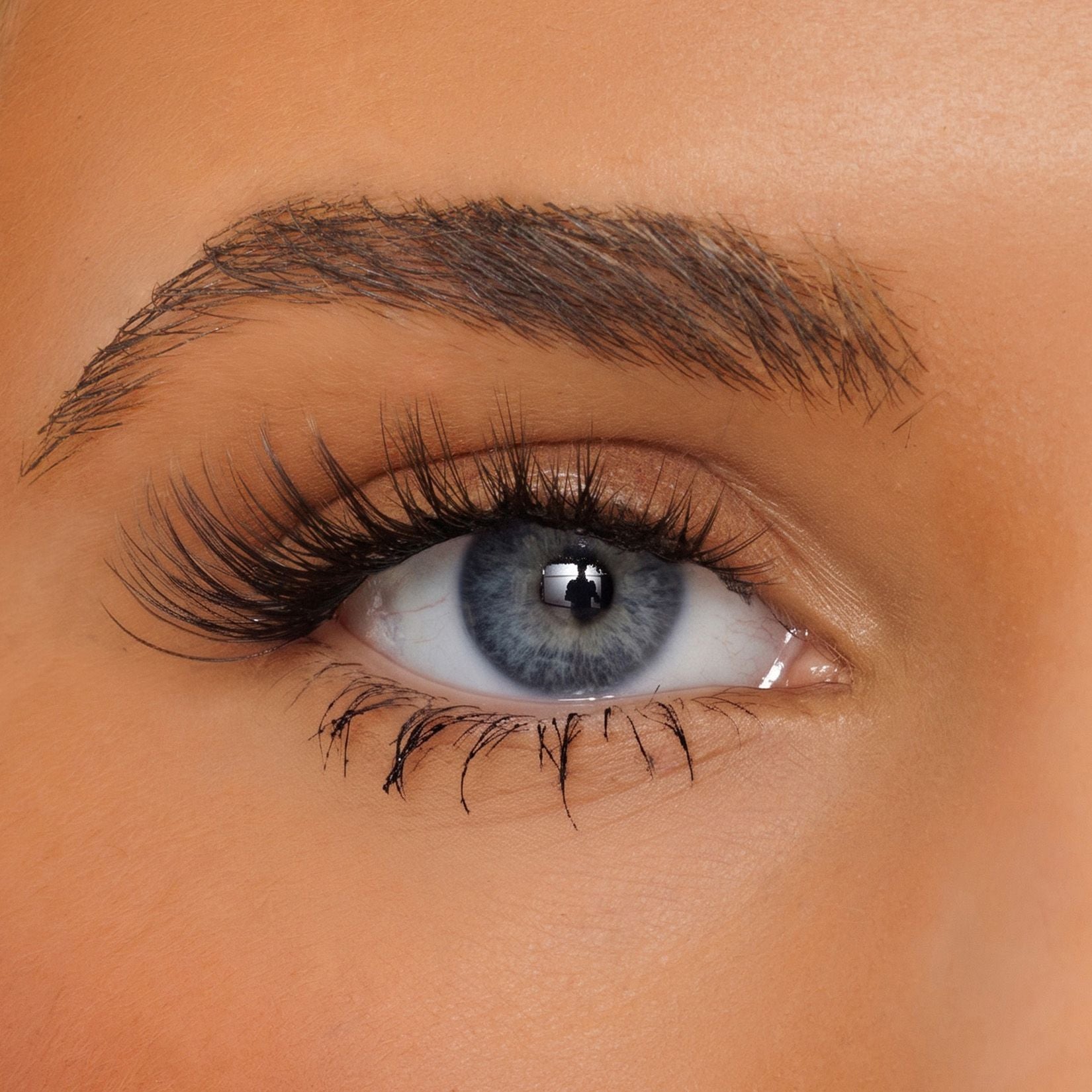 Close-up of a person's eye with long, dark, curled false eyelashes and a neatly shaped dark brown eyebrow on warm tan skin.